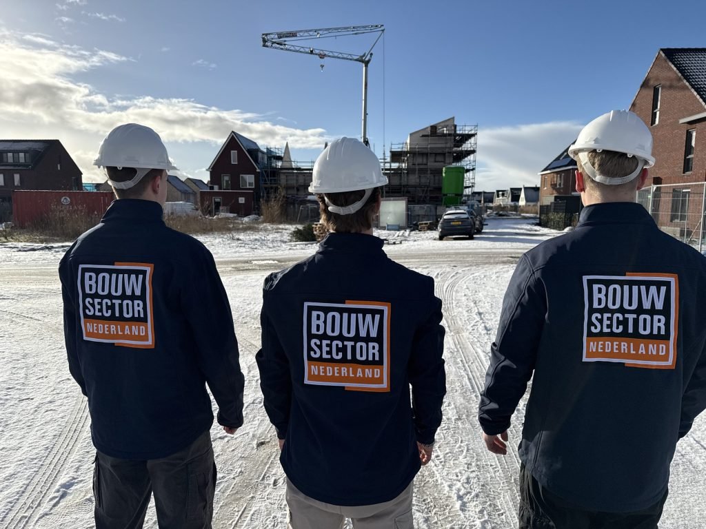 Drie vakmensen met witte helm en het logo op de rug kijken naar huizen in aanbouw in de sneeuw, met een bouwkraan erachter